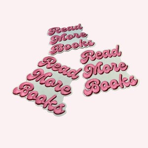 Op de afbeelding: Drie geborduurde patches met de tekst Read More Books in een cursief roze lettertype op een witte achtergrond. De patches zijn gevormd om de contouren van de letters te volgen, wat zorgt voor een duidelijk en leesbaar ontwerp per stuk.