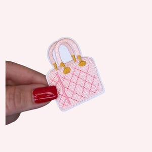 May include: A pink embroidered patch in the shape of a handbag. The patch features a quilted design with gold-colored accents on the handles. The patch is held against a light pink background.