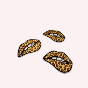 May include: Three embroidered patches shaped like lips, each featuring a brown and tan leopard print pattern. The lips are depicted with a slightly open mouth, revealing white teeth and a dark outline, creating a distinct graphic design element.