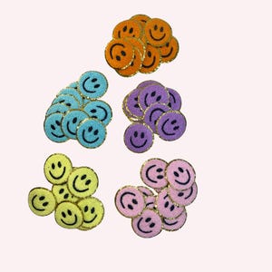 Peut inclure: Plusieurs écussons brodés en forme de smiley, de couleurs orange, bleu, violet, jaune et rose. Chaque écusson présente un sourire noir cousu et une bordure dorée. Parfaits pour personnaliser vêtements et accessoires.