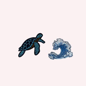 Può includere: Due toppe ricamate: una tartaruga marina blu scuro con dettagli blu chiaro e un'onda oceanica stilizzata blu e bianca. Le toppe sono probabilmente per abbigliamento o accessori.