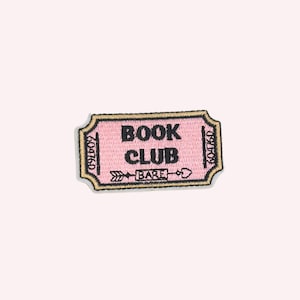 Puede incluir: Un parche bordado rectangular con forma de boleto, fondo rosa claro y borde oscuro. El texto dice BOOK CLUB en negro, con un gráfico de flecha que apunta a una pequeña caja que contiene la palabra BARE y un símbolo de corazón.