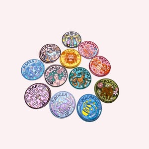 May include: A collection of twelve colorful, round zodiac sign patches. Each patch features a unique illustration and the name of the astrological sign in a decorative font. The patches are arranged on a light pink background.