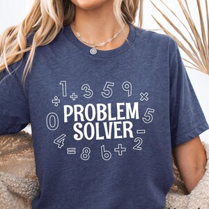 May include: Heather navy blue t-shirt with the text "PROBLEM SOLVER" surrounded by math symbols and numbers. The shirt is a short-sleeved crew neck.