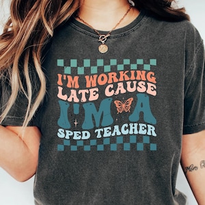 May include: Dark gray t-shirt with the text "I'M WORKING LATE CAUSE I'M A SPED TEACHER" in a retro font. The design includes a butterfly and a checkered pattern in teal and orange. The shirt is a comfortable, casual style.