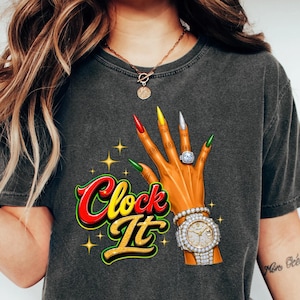 May include: A charcoal gray t-shirt featuring a graphic of a hand with long, multi-colored nails, a diamond ring, and a diamond-encrusted watch. The text "Clock It" is printed in a bold, stylized red and yellow font with small star accents.