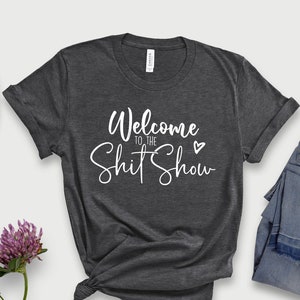May include: Dark grey heather t-shirt with the text "Welcome to the Shit Show" in white cursive lettering with a small heart.