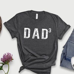 May include: Dark grey heather t-shirt with the text "DAD³" in white. The shirt is laid flat on a white background with a pair of jeans and sneakers.