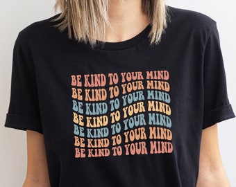 May include: Black t-shirt with the text "BE KIND TO YOUR MIND" in a retro, wavy font. The text is in a rainbow of colors, including orange, yellow, blue, and pink. The shirt has short sleeves and a relaxed fit.