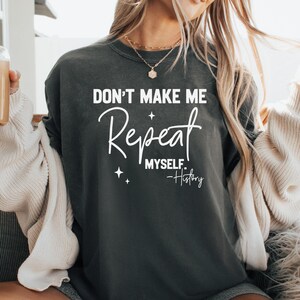 May include: A dark gray t-shirt with the text "Don't Make Me Repeat Myself. -History" in white lettering. The shirt features a cursive font and small star accents.