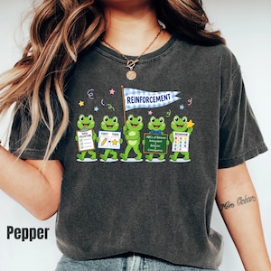 May include: A charcoal gray t-shirt featuring a graphic of five green frogs. One frog holds a blue pennant that says REINFORCEMENT. Other frogs hold signs labeled DATA COLLECTION, FIRST THEN, ABCs of Behavior, and BEHAVIOR with colorful stars.