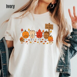 May include: An ivory t-shirt featuring a graphic of a pumpkin, a mug, a maple leaf, and an acorn with faces. The text reads "SENSORY REGULATION," "FINE MOTOR SKILLS," "DAILY ACTIVITIES," "CONFIDENCE & INDEPENDENCE," and "OT HELPS YOU GROW" on a sign.