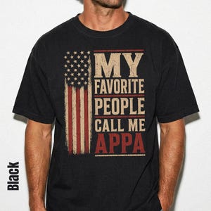 May include: A black short-sleeved t-shirt featuring a distressed graphic of the American flag alongside the text "MY FAVORITE PEOPLE CALL ME APPA" in a bold, weathered font. The design uses a muted color palette of off-white and faded red.