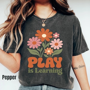May include: A charcoal gray short-sleeve t-shirt featuring a graphic of orange and pink flowers with green leaves. Below the floral design, the text reads "PLAY is Learning" in orange and green lettering. The shirt has a crew neckline.