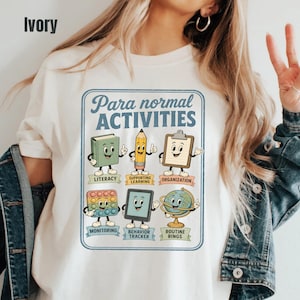 May include: An ivory t-shirt featuring a graphic with the text Para normal ACTIVITIES and six cartoon characters representing school supplies with labels: LITERACY, SUPPORTING LEARNING, ORGANIZATION, MONITORING, BEHAVIOR TRACKER, and ROUTINE RINGS.