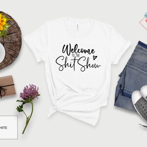 May include: White t-shirt with the text "Welcome to the Shit Show" in black cursive lettering with a small heart symbol.