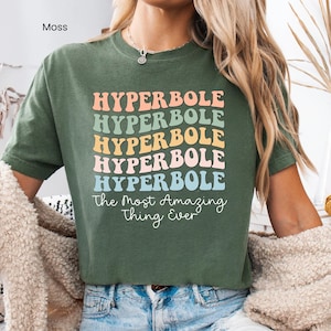 May include: A moss green t-shirt with the word "HYPERBOLE" in a stacked, colorful design. Below the word is the phrase "The Most Amazing Thing Ever" in a cursive font. The shirt is short-sleeved and made of a soft material.
