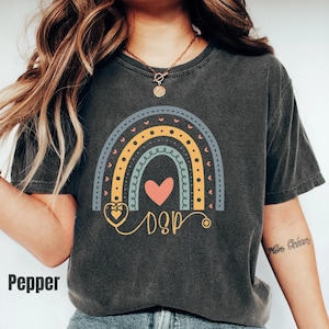 May include: A charcoal gray short-sleeve t-shirt featuring a graphic of a multi-colored rainbow with a central pink heart and a stethoscope design. The text DSP is printed in a cursive font below the heart. The shirt has a crew neckline.