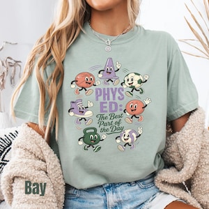 May include: A sage green t-shirt featuring a graphic print of cartoon sports equipment including a basketball, soccer ball, volleyball, kettlebell, sneaker, and traffic cone. The text reads "PHYS ED: The Best Part of the Day" in purple and black.