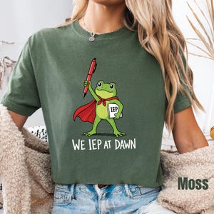May include: A moss green t-shirt featuring a graphic of a green frog wearing a red cape, holding a red pen and a white folder labeled IEP. Below the frog, the white text reads WE IEP AT DAWN. The shirt has a crew neckline and short sleeves.