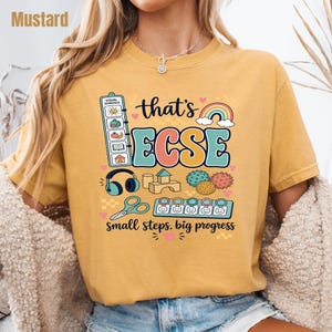 May include: A mustard yellow t-shirt featuring a graphic print with the text "that's ECSE" and "small steps, big progress." The design includes a visual schedule, a rainbow, headphones, wooden blocks, sensory balls, scissors, and a token board.