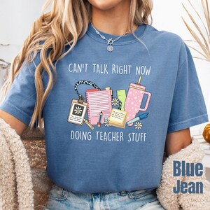May include: A blue t-shirt featuring a graphic print with the text CAN'T TALK RIGHT NOW DOING TEACHER STUFF. The design includes a pink tumbler, a notebook, a lanyard, a pencil, a pen, and a notepad, all decorated with small white daisy flowers.