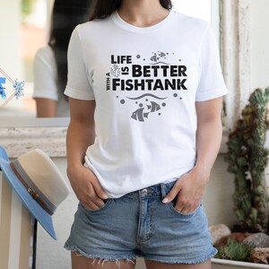 Puede incluir: Camiseta blanca con el texto negro "LIFE IS BETTER WITH A FISHTANK". El dise&ntilde;o incluye peces y burbujas. La persona lleva pantalones cortos de mezclilla. Un sombrero azul y beige est&aacute; en una silla en el fondo.