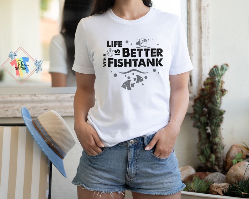 Puede incluir: Camiseta blanca con el texto negro "LIFE IS BETTER WITH A FISHTANK". El dise&ntilde;o incluye peces y burbujas. La persona lleva pantalones cortos de mezclilla. Un sombrero azul y beige est&aacute; en una silla en el fondo.