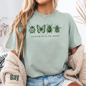 May include: A sage green short-sleeve t-shirt featuring a dark green graphic of a ladybug, butterfly, beetle, and bee arranged in a row. Below the insects, the text reads "learning is in our nature" in a simple, dark green lowercase font.
