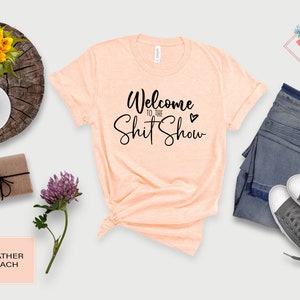 May include: A peach colored t-shirt with the text "Welcome to the Shit Show" in black cursive lettering with a small black heart.