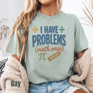 May include: A sage green short-sleeve t-shirt featuring the text I HAVE PROBLEMS (math ones) in blue and orange. The design includes a yellow plus sign, a blue division symbol, a green pi symbol, and a brown ruler graphic on the front.