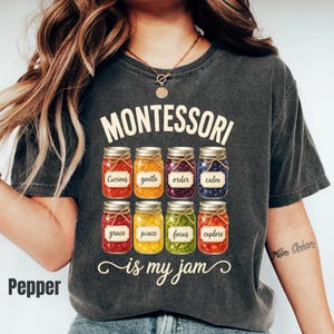 May include: A charcoal gray t-shirt featuring the text "MONTESSORI is my jam" in cream lettering. Below the text are eight glass jars filled with colorful items, each labeled with words including curious, gentle, order, calm, grace, peace, focus, and explore.