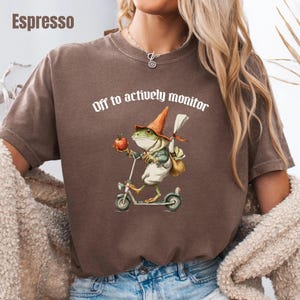 May include: An espresso brown short-sleeve t-shirt featuring a graphic of a green frog wearing an orange pointed hat, riding a scooter, and holding a red apple. The text above the frog reads, "Off to actively monitor" in white lettering.