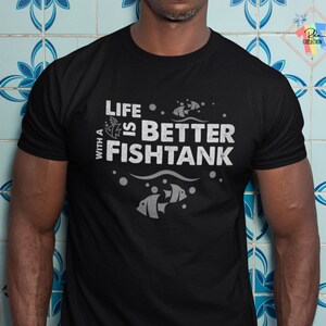 Puede incluir: Camiseta negra con el texto blanco "LIFE IS BETTER WITH A FISHTANK". El dise&ntilde;o incluye ilustraciones de peces, burbujas y olas. La camiseta es de cuello redondo y tiene mangas cortas.