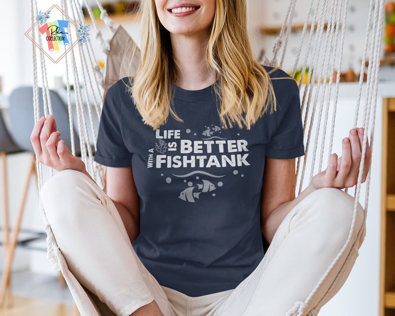 Puede incluir: Camiseta azul marino con el texto blanco "Life is Better with a Fishtank". La camiseta presenta ilustraciones de peces. Es una prenda informal.