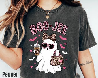 Può includere: Una t-shirt grigio antracite con la grafica di un fantasma bianco che indossa occhiali da sole a forma di cuore e un fiocco leopardato. Il fantasma tiene in mano un bicchiere e una borsa leopardati. Il testo BOO-JEE è stampato in rosa leopardato.