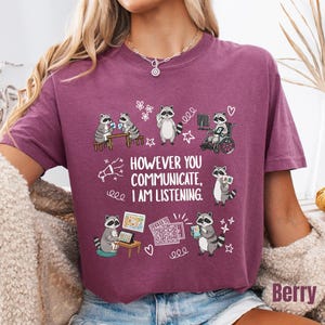 May include: A berry-colored t-shirt featuring a graphic of raccoons using various communication methods. The text reads, "HOWEVER YOU COMMUNICATE, I AM LISTENING." The design includes illustrations of raccoons with tablets, a wheelchair, and communication boards, surrounded by small white line-art doodles of hearts, stars, and flowers.