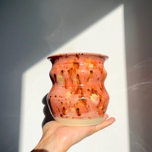 Op de afbeelding: Een keramieken beker met een golvend, zandloperachtig silhouet, voorzien van een glanzend glazuur in koraalroze en oranje tinten met donkerbruine druppelachtige spikkels en lichtere gele vlekken, afgewerkt met een beige, ongeglazuurde voet.