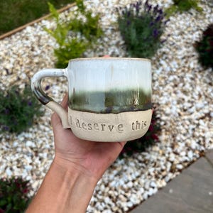 May include: A ceramic mug with a two-tone glaze featuring a glossy white top and a dark green middle section, transitioning to a matte beige base. The text "I deserve this" is embossed in lowercase letters on the lower beige portion of the mug.