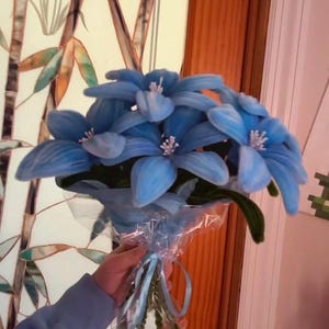 May include: A bouquet of light blue artificial flowers with white centers, wrapped in clear cellophane and tied with a light blue ribbon. The flowers are held in a person's hand. The background includes a stained glass window and a wooden door.