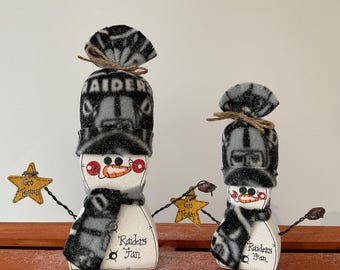 Puede incluir: Dos figuras de muñeco de nieve de madera blanca con gorros y bufandas de forro polar negro y gris con la marca Raiders. Cada muñeco tiene una nariz de zanahoria naranja, mejillas rojas y el texto Raiders Fan. Sostienen estrellas amarillas.