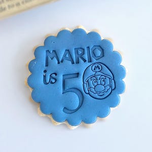 Puede incluir: Una galleta azul con borde festoneado, con el texto "MARIO is 5" y una imagen de dibujos animados de Mario. La galleta tiene un borde marrón claro, con un paquete de galletas en el fondo.