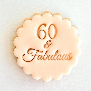 May include: A round, scalloped-edge biscuit with a smooth, pale peach-coloured icing finish. The centre features embossed text that reads 60 & Fabulous in a combination of bold block numerals and an elegant, flowing cursive script font.
