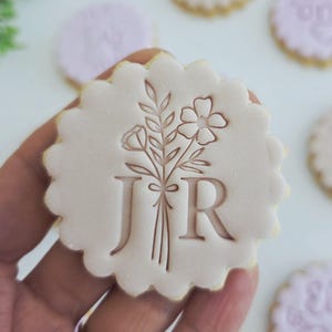 May include: A hand holds a light pink, scalloped-edge cookie with a floral design and the letters "J R". Other cookies with floral and text designs are in the background. These cookies are likely for a special event or celebration.