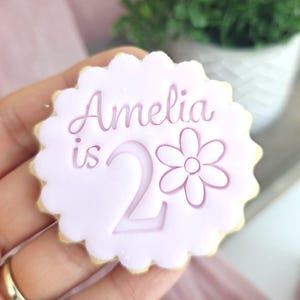 May include: A light purple cookie with a scalloped edge. The cookie has the name "Amelia is 2" and a flower design. The cookie is held in a hand, with a gold ring visible. A green plant in a white pot is in the background.