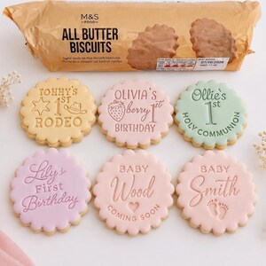 May include: Six round, scalloped-edge biscuits with pastel icing and embossed text. Designs include Tommy's 1st Rodeo, Olivia's Berry 1st Birthday, Ollie's 1st Holy Communion, Lily's First Birthday, Baby Wood Coming Soon, and Baby Smith with footprints.
