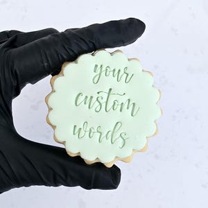 May include: A custom cookie with light green icing and a scalloped edge. The words "your custom words" are written in a cursive font on the icing. The cookie is held by a gloved hand.
