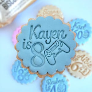 Peut inclure: Une pile de biscuits décorés avec un glaçage bleu clair. Le biscuit du dessus porte le nom "Kayen", le chiffre "8" et un motif de manette de jeu vidéo. D'autres biscuits portent des noms et des chiffres.