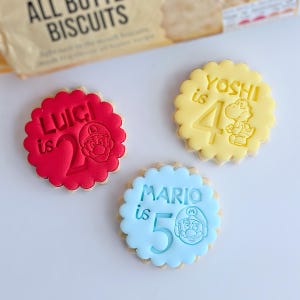 May include: Three decorated sugar biscuits with scalloped edges. One red biscuit says "Luigi is 2" with a Mario character. A yellow biscuit says "Yoshi is 4" with a Yoshi character. A blue biscuit says "Mario is 5" with a Mario character.