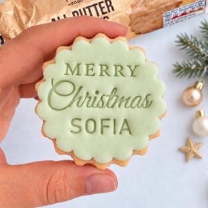 May include: A round scalloped biscuit with light green icing featuring the embossed text MERRY Christmas SOFIA in a dark green font. The biscuit has a golden brown edge and is held against a background with a blurred food package and ornaments.